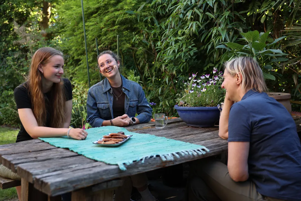 Drie jonge vrouwen lachen samen aan een houten tuintafel